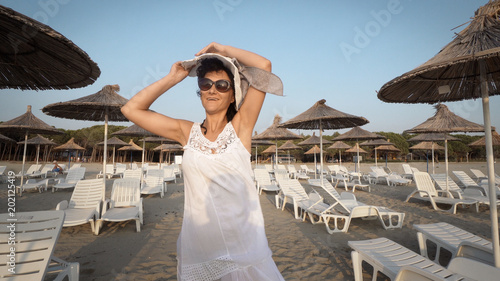 Sexy woman in white dress and hat walking on empty beautiful beach with straw umbrellas and beds