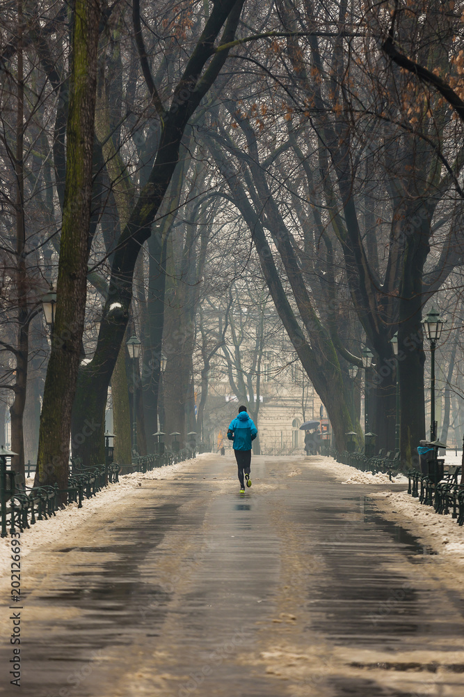 Man running along path surrounded by trees, in winter, using blue sports jacket, Krakow, Poland