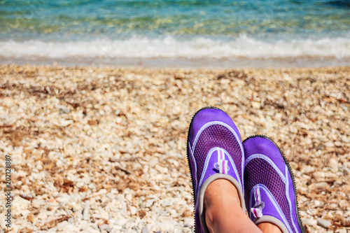 Female legs with Swimming neoprene shoes.Water shoes, swimming shoe in purple neoprene on rocks in water on beach. Coral slippers on rocky beach