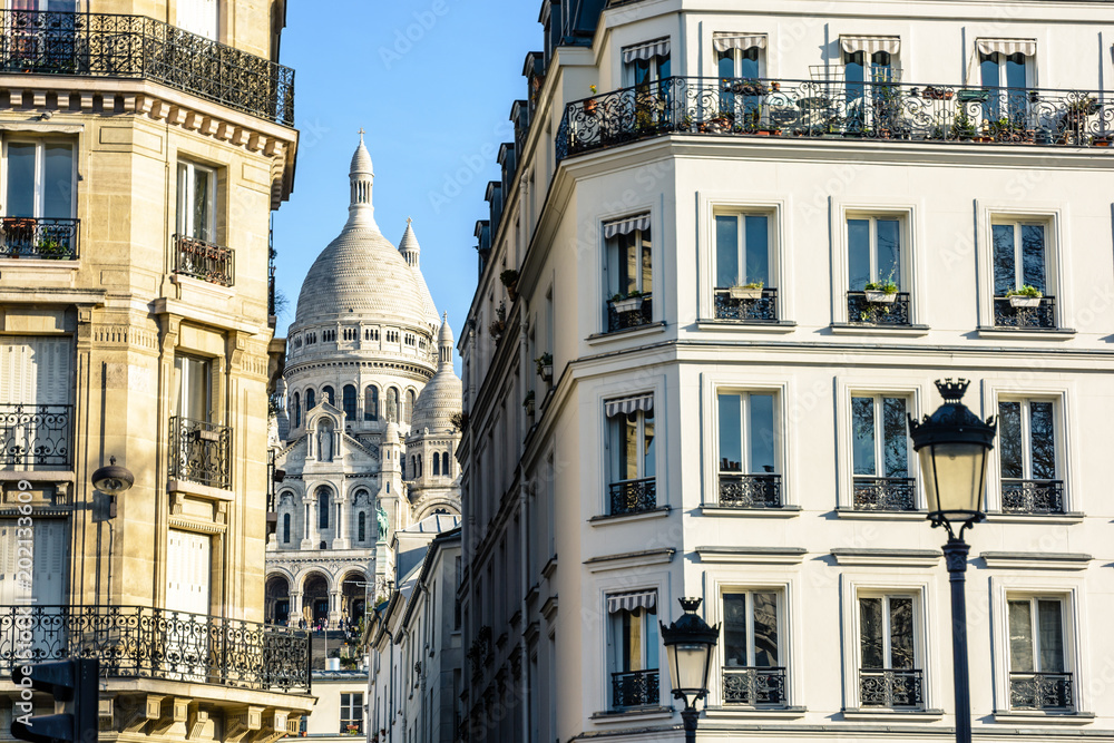 The Basilica of the Sacred Heart in Paris seen through a narrow street between typical buildings with vintage street lights in the foreground under a clear blue sky.