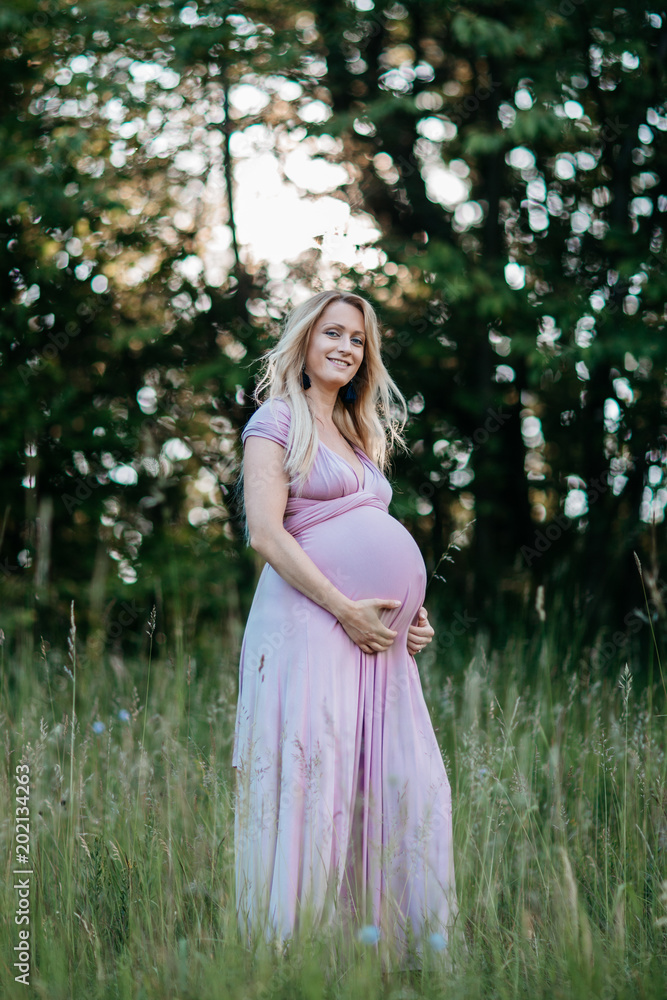 A happy pregnant woman in a summer dress touching her belly. A portrait of a smiling expecting mother stroking her pregnancy bump.