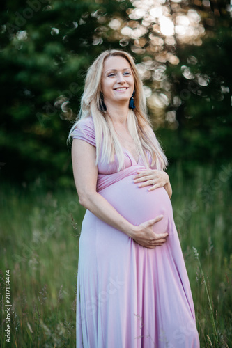 A young expectant mother gently touching her pregnancy belly. A portrait of a smiling pregnant woman enjoying being in the nature on a summer evening.