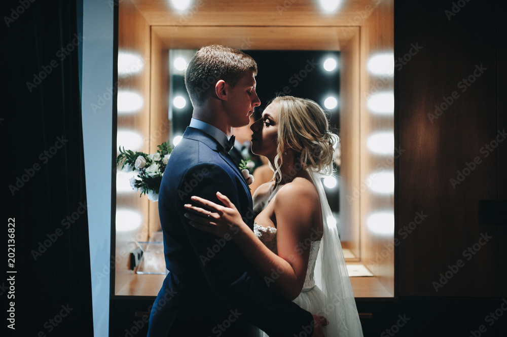 Cute newlyweds hug and are reflected in a mirror with lamps and herds. Portrait of a newlywed couple in a modern studio. Film effect. Cute newlyweds hug and are reflected in a mirror with lamps and herds. Portrait of a newlywed couple in a modern studio. Film effect.