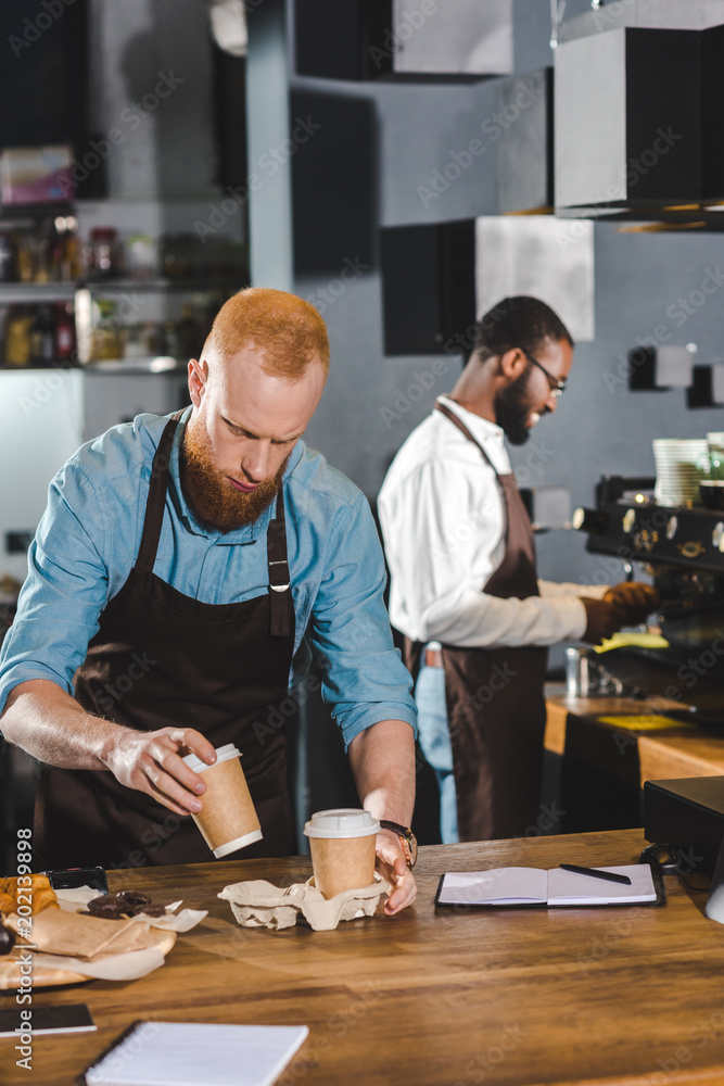 Fototapeta premium smiling african american male barista using coffee machine and colleague putting paper cups of coffee in cardboard