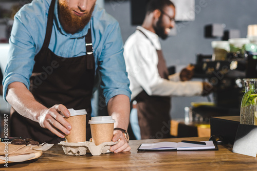 cropped shot of male barista in apron putting paper cups of coffee in cardboard and smiling colleague using coffee machine behind