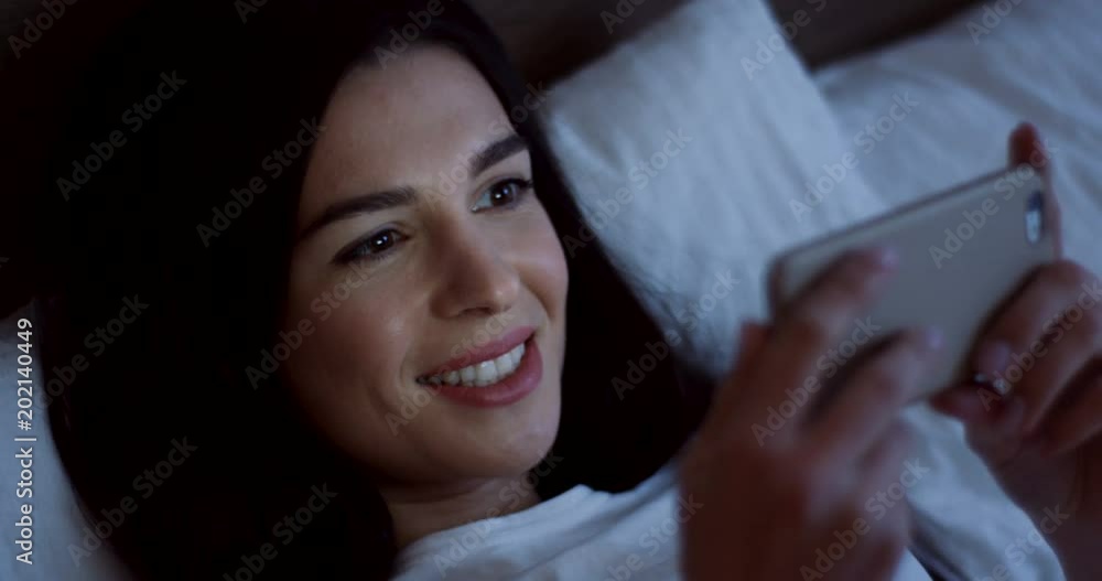 Close up of the attractive smiled brunette woman with long black hair lying in the bed and watching a video on the smartphone late in the evening. Indoor Close up of the attractive smiled brunette woman with long black hair lying in the bed and watching a video on the smartphone late in the evening. Indoor