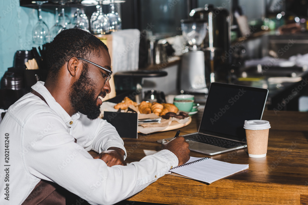 smiling young african american barista in eyeglasses taking notes and using laptop with blank screen in cafe smiling young african american barista in eyeglasses taking notes and using laptop with blank screen in cafe