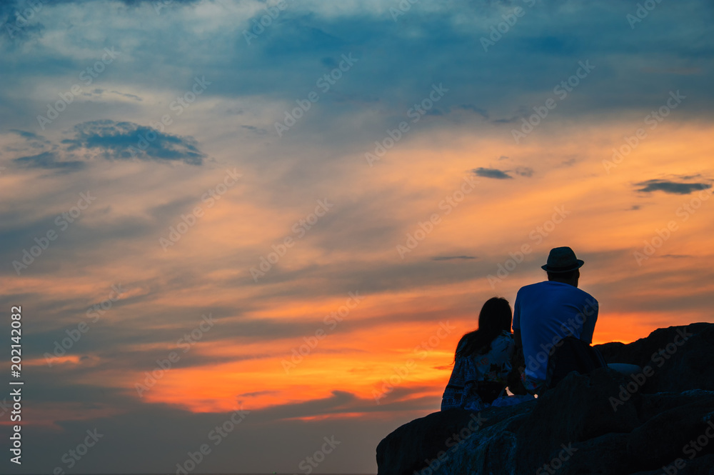 Silhouette of a loving couple sitting on a rock over evening tropical sea, over blurred background. Sunset pastel background with light fair. Silhouette of a loving couple sitting on a rock over evening tropical sea, over blurred background. Sunset pastel background with light fair.