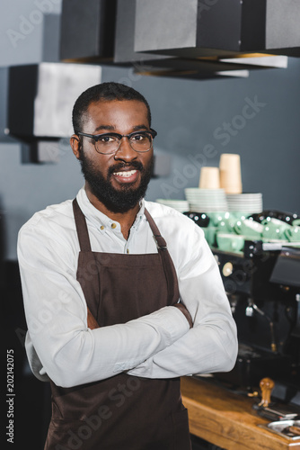 confident african american barista in eyeglasses standing with crossed arms and smiling at camera in coffee shop