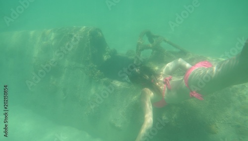Billede på lærred UNDERWATER: Intrigued woman in pink bikini looks closely at sunken aircraft