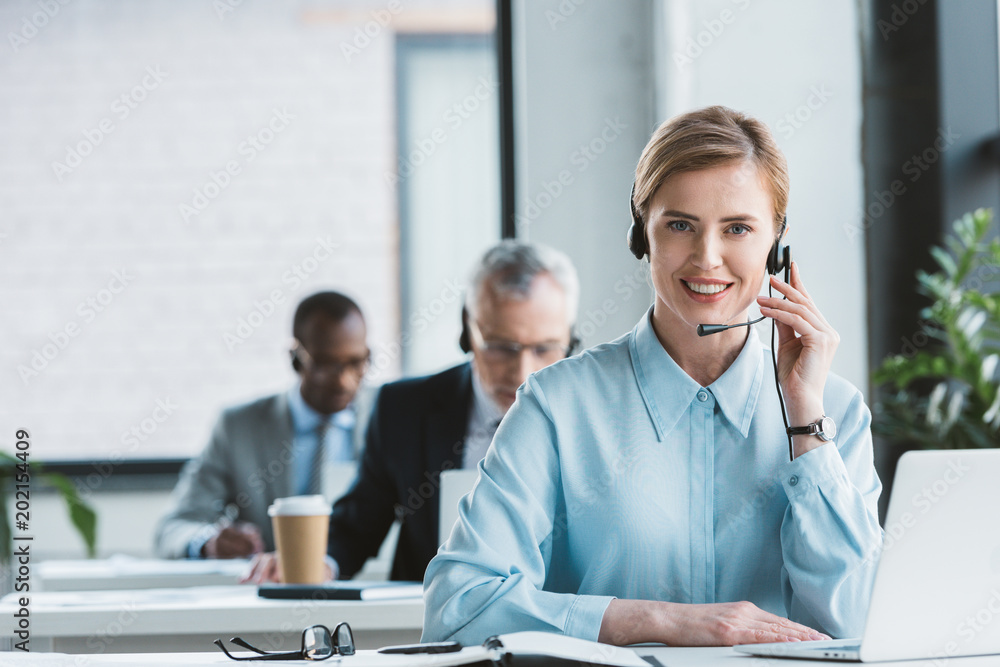 attractive businesswoman in headset using laptop and smiling at camera attractive businesswoman in headset using laptop and smiling at camera