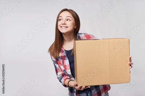 Delivery, relocation and unpacking. Smiling young woman holding cardboard box, looking at blank copy space to the side, isolated