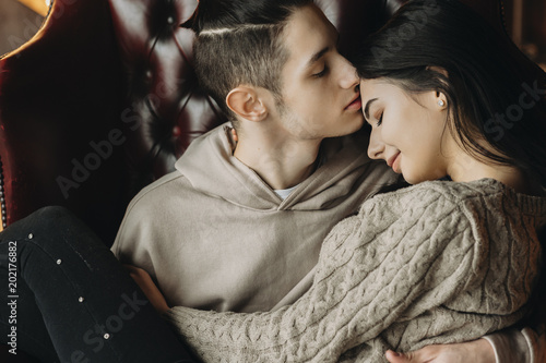 Cuadro en lienzo Close up portrait of a beautiful young couple sitting on a leather chair where boy is kissing his girlfriend on the forehead while she is smiling with closed eyes