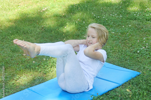 Beautiful blonde preteen girl in light clothing practicing yoga on a mat in the park. Healthy lifestyle. Outdoors workout.