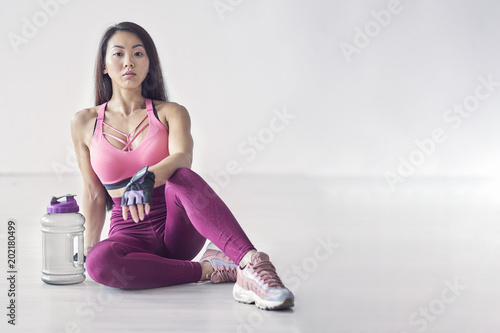 Sport woman sitting and resting after workout or exercise in fitness gym with protein shake or drinking water on floor. Relax concept. Strength training and Body build up theme. Copy space.