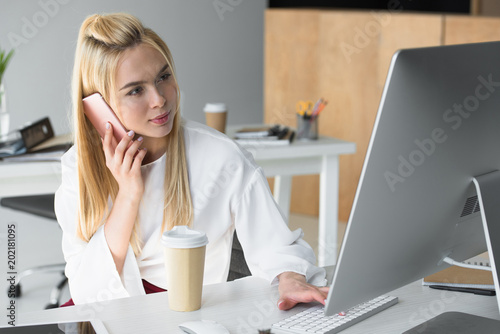 beautiful young businesswoman talking by smartphone and using desktop computer in office