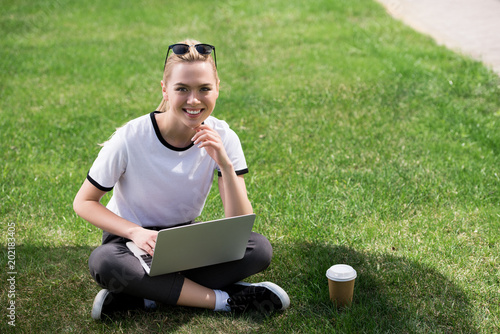 beautiful blonde girl smiling at camera while using laptop on grass