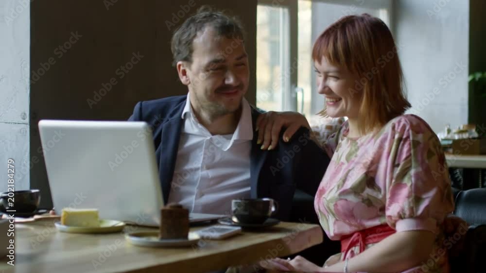 Medium shot of happy young woman in floral dress and cheerful man in jacket sitting at tablet in cafe and joking while talking to someone on video call via laptop Medium shot of happy young woman in floral dress and cheerful man in jacket sitting at tablet in cafe and joking while talking to someone on video call via laptop