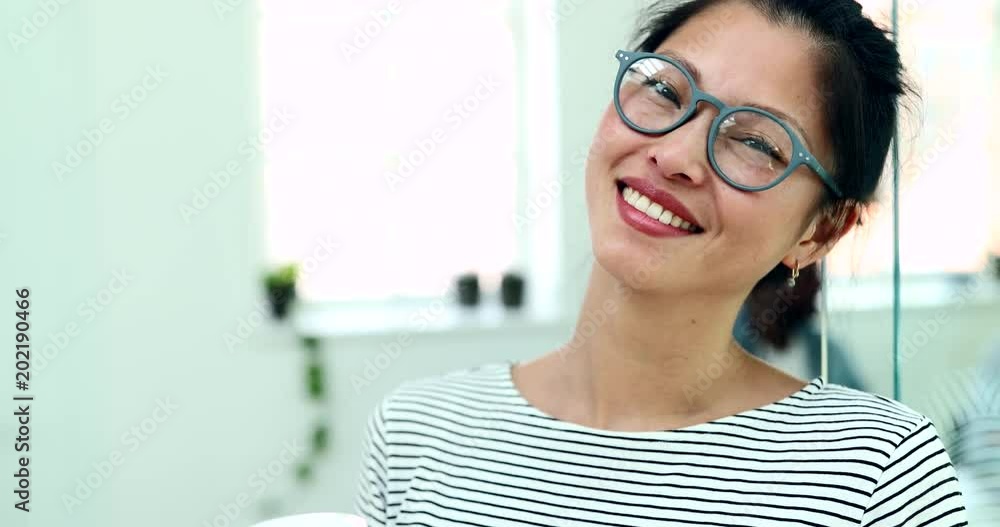 Smiling young Asian businesswoman wearing glasses and drinking a coffee while standing in an office with colleagues at work in the background Smiling young Asian businesswoman wearing glasses and drinking a coffee while standing in an office with colleagues at work in the background