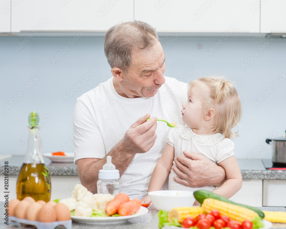 Senior man feeding baby girl with a spoon at kitchen