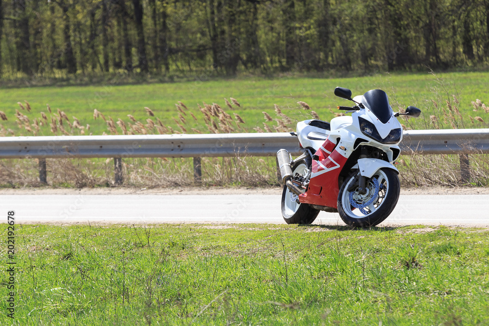 Obraz premium A motorcycle racer stands by the side of an empty road on a sunny day. A lot of green grass on the background. Nobody