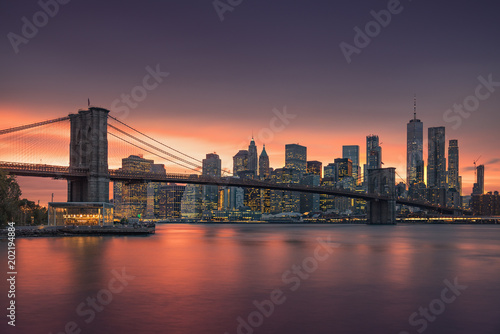 Famous Brooklyn Bridge in New York City with financial district - downtown Manhattan in background. Sightseeing boat on the East River and beautiful sunset over Jane's Carousel.
