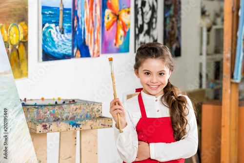 kid holding painting brush and looking at camera in workshop of art school