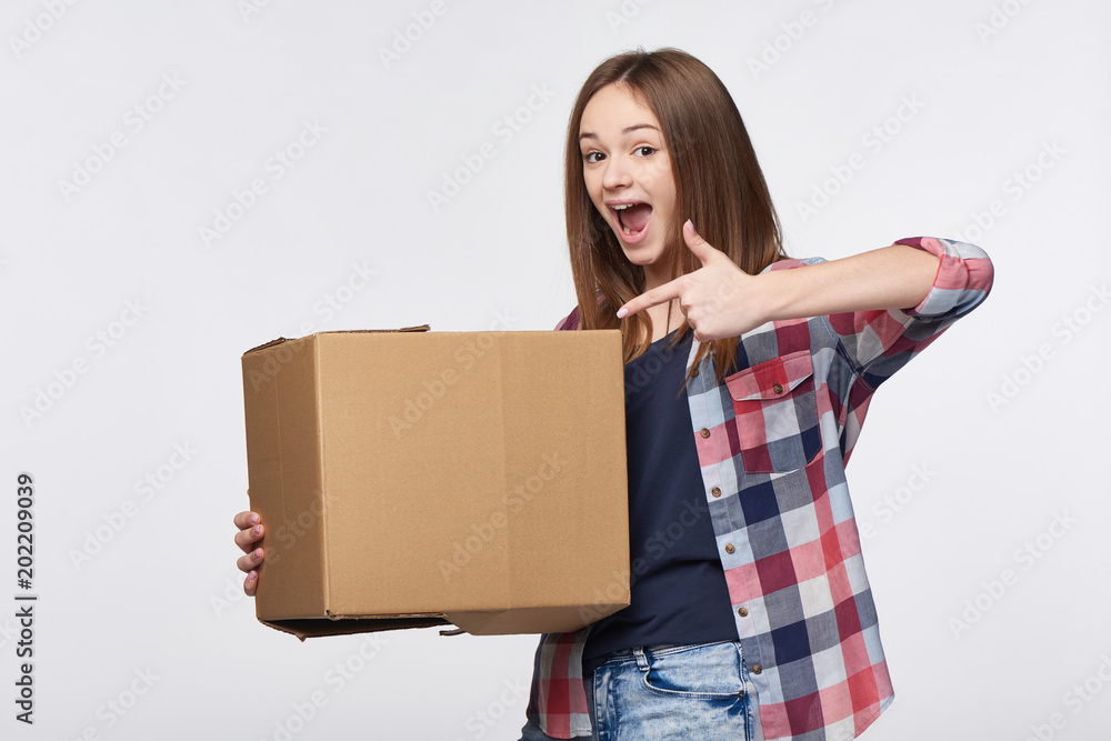 Delivery, relocation and unpacking. Happy excited young woman holding cardboard box pointing emotionally at box Delivery, relocation and unpacking. Happy excited young woman holding cardboard box pointing emotionally at box