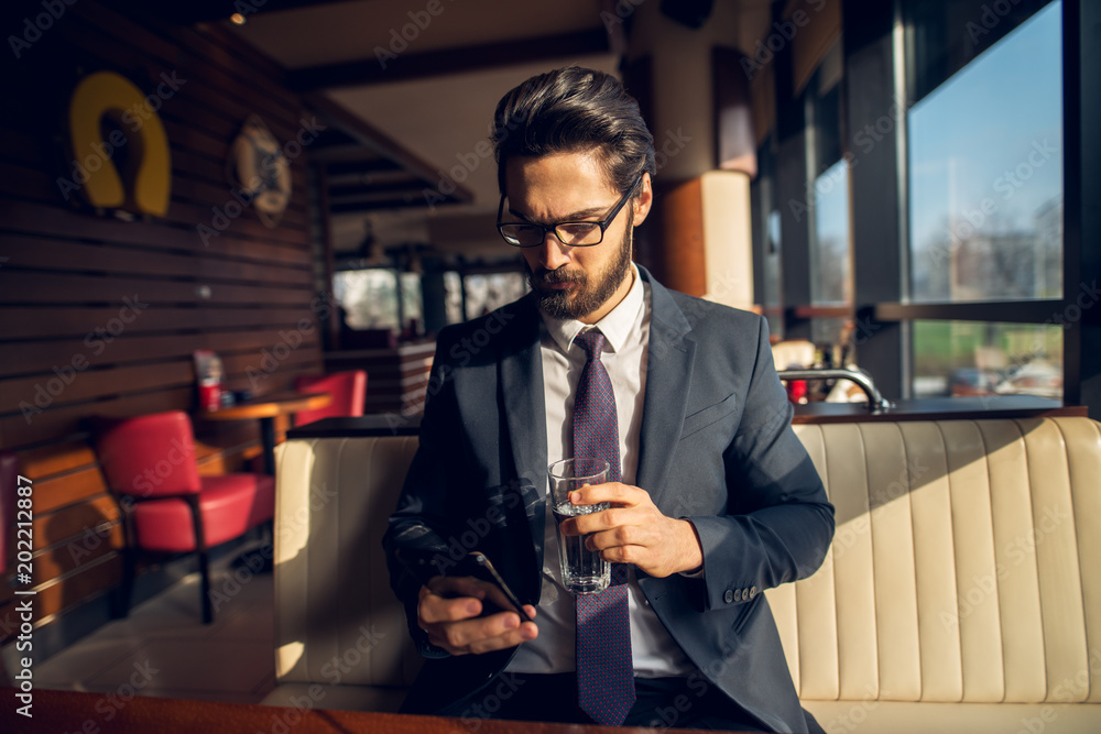 Obraz premium Successful stylish focused handsome bearded businessman in the suit looking at a mobile while holding a glass of water in a cafe or restaurant.