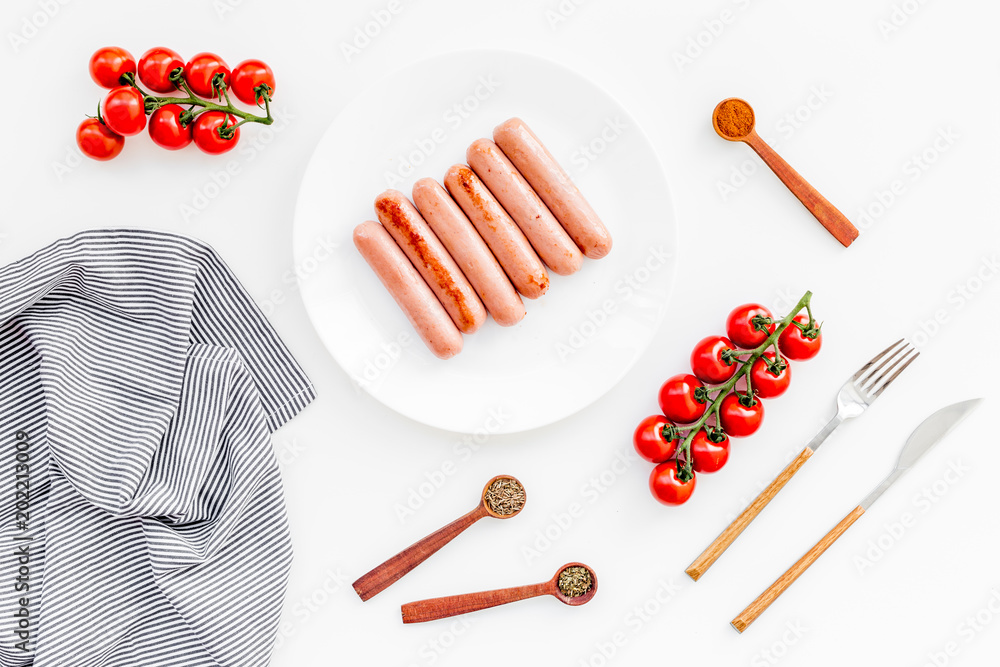Grilled sausages on white table near cherry tomatoes and spices in wooden spoons top view Grilled sausages on white table near cherry tomatoes and spices in wooden spoons top view