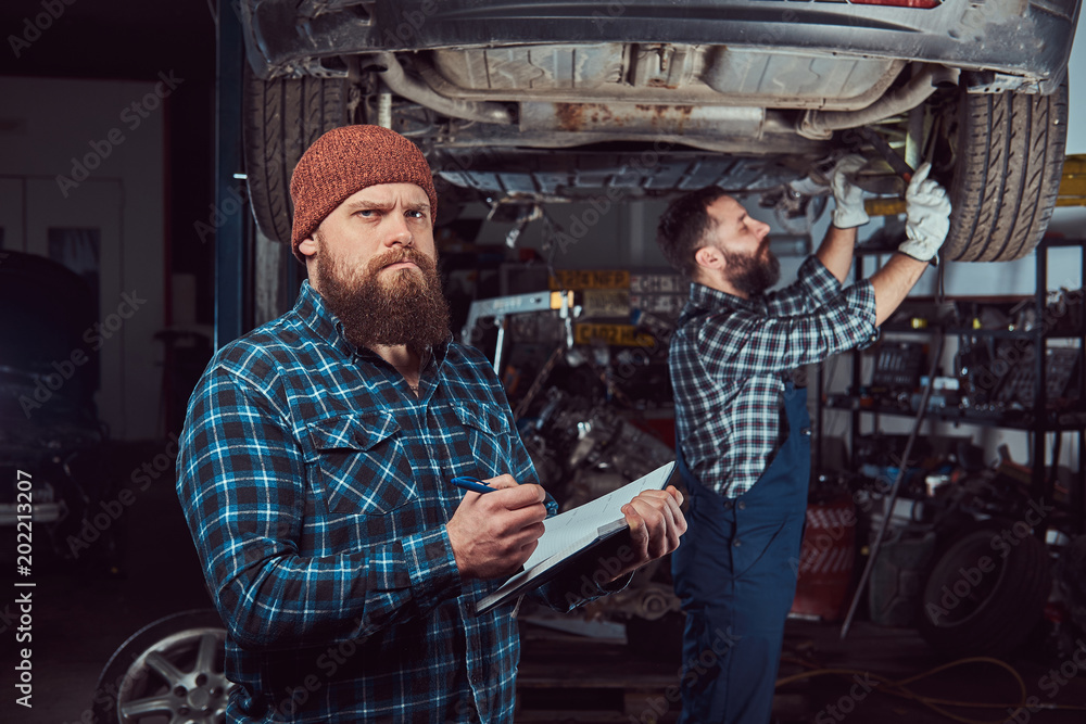 Two bearded brutal mechanics repair a car on a lift in the garage. Two bearded brutal mechanics repair a car on a lift in the garage.