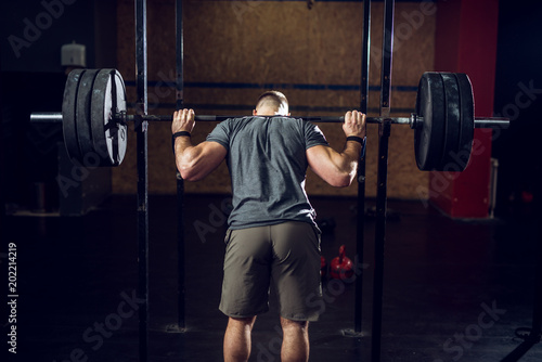 Rear view of young short hair focused strong muscular bodybuilder man standing with a heavy barbell behind the neck while preparing for squat exercise.