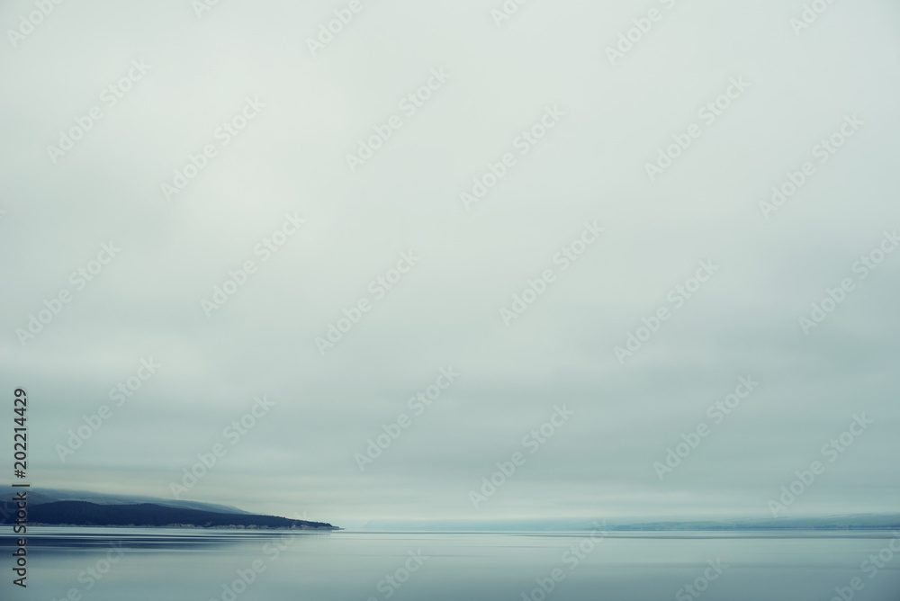 Paisaje azul minimalista de un lago tranquilo, un cielo denso blanco y pequeñas montañas distantes casi invisibles en un día nublado de niebla.