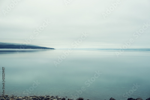 Paisaje azul minimalista de un lago tranquilo, un cielo denso blanco y pequeñas montañas distantes casi invisibles en un día nublado de niebla.