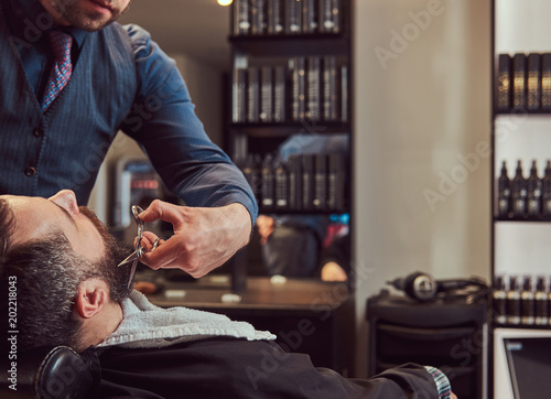 Professional hairdresser modeling beard with scissors and comb at the barbershop.