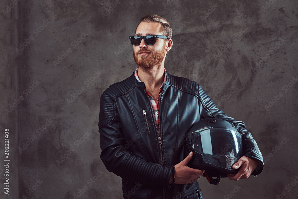 Plakat Portrait of a handsome stylish redhead biker in a black leather jacket and sunglasses, holds motorcycle helmet, posing in a studio.