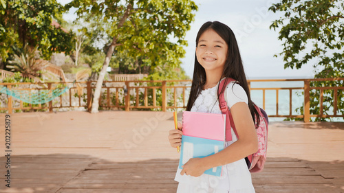 A charming philippine schoolgirl with a backpack and books in a park off the coast. A girl joyfully poses, raising her hands up with textbooks in her hands. Warm sunny day. photo