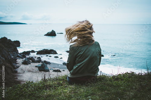 Φωτογραφία girl sitting on a cliff overlooking the ocean with her hair blowing in the wind