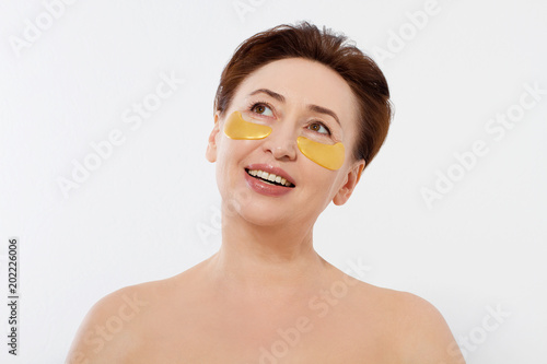 Beauty portrait of middle age woman with wrinkles and a gold patch under eye isolated on white background. Collagen mask and spa concept. Copy space. Summer skin care