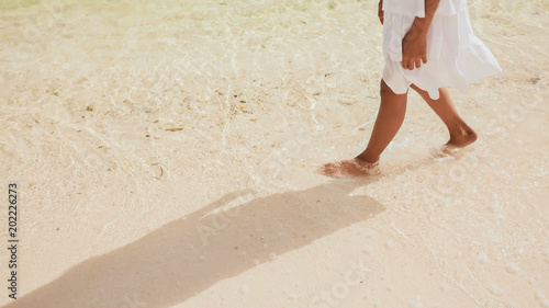 The naked and tanned legs of an philippine schoolgirl in a white dress walk on white sand, touching the foamy waves by the shore. Tropical landscape. Childhood.