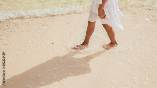 The tanned legs of an philippine schoolgirl in a white dress run along white sand, touching the waves of foam on the beach. Tropical landscape. Childhood.