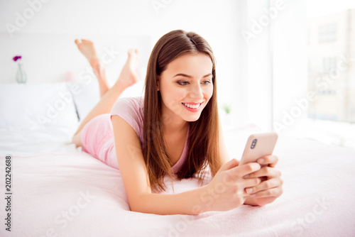 Portrait of carefree, pretty, charming, cheerful, pretty, cute girl having smart phone in hands lying on stomach chatting with friends, checking email, texting sms, using wifi, 5G internet