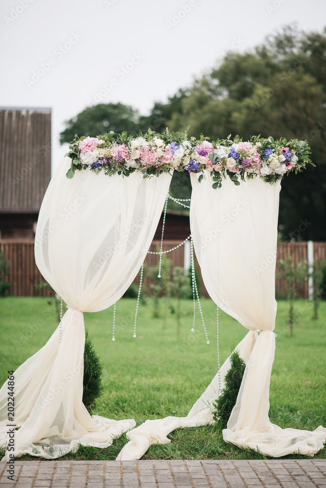 Wedding arch decorated with cloth and flowers outdoors. Beautiful wedding set up. Wedding ceremony on green lawn in the garden. Part of the festive decor, floral arrangement.