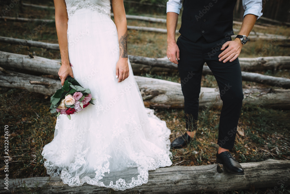 The groom in a stylish tuxedo and the bride in a wedding dress with a bouquet in hands stand near a log in the open air