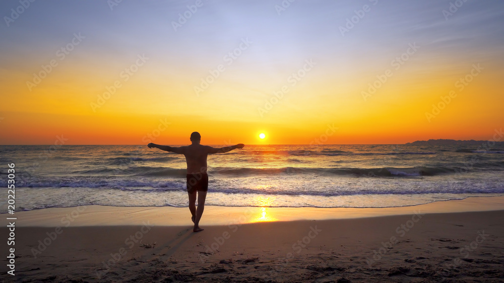 Man with hands wide open in VICTORY pose walks on empty ocean beach into water with vibrant sunset sun at background, cinematic steadicam shot
