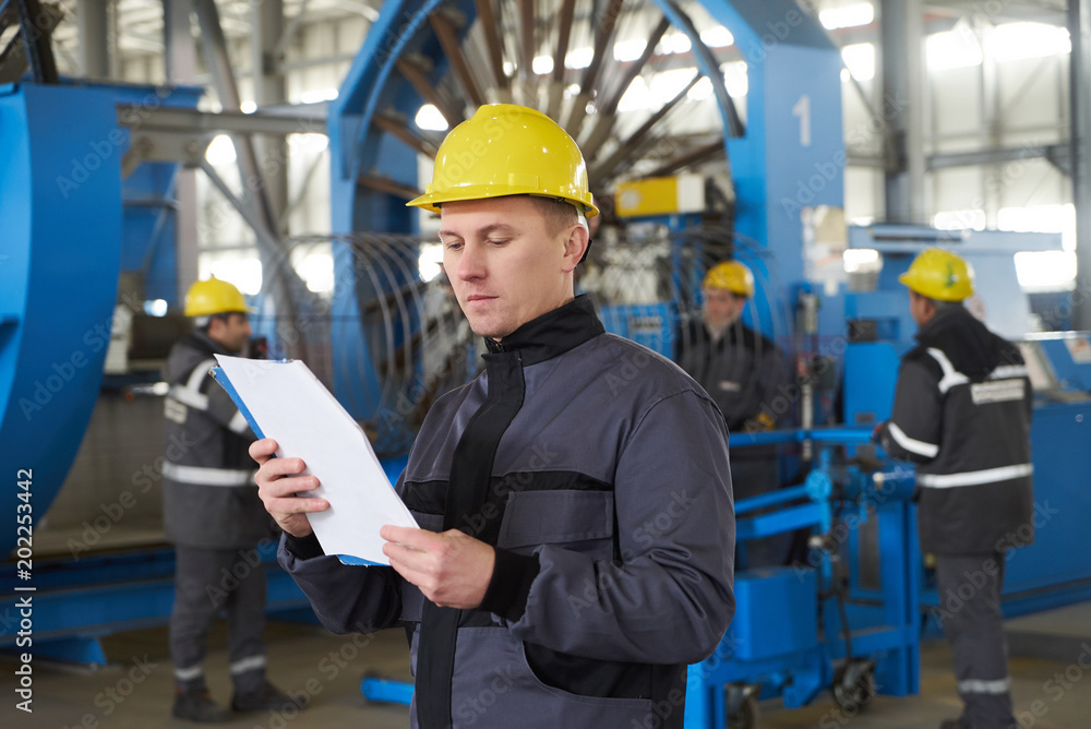 Portrait of factory worker holding paper clipboard. Young engineer in uniform and hardhat taking notes working at metal production factory, copy space Portrait of factory worker holding paper clipboard. Young engineer in uniform and hardhat taking notes working at metal production factory, copy space