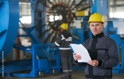Young Worker reading paper documents while standing at factory with working machines on background. 