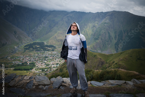 Young man stands on the top and relaxes. Traveler in casual clothes on background of small city in the valley and great mountains.