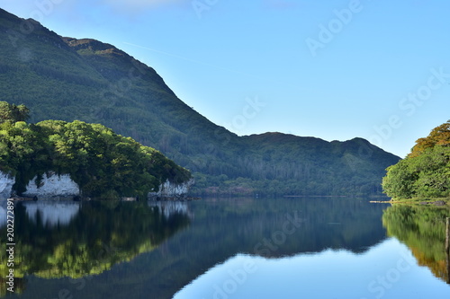 Reflections of hills forests and rock formations on calm surface of Muckross Lake in Ireland.