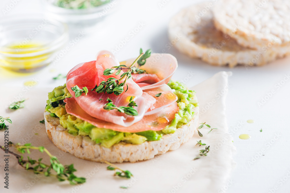 Crisp bread healthy snack with Parma ham, avocado spread, olive oil, thyme. Easy breakfast close-up on a white background with copy space.